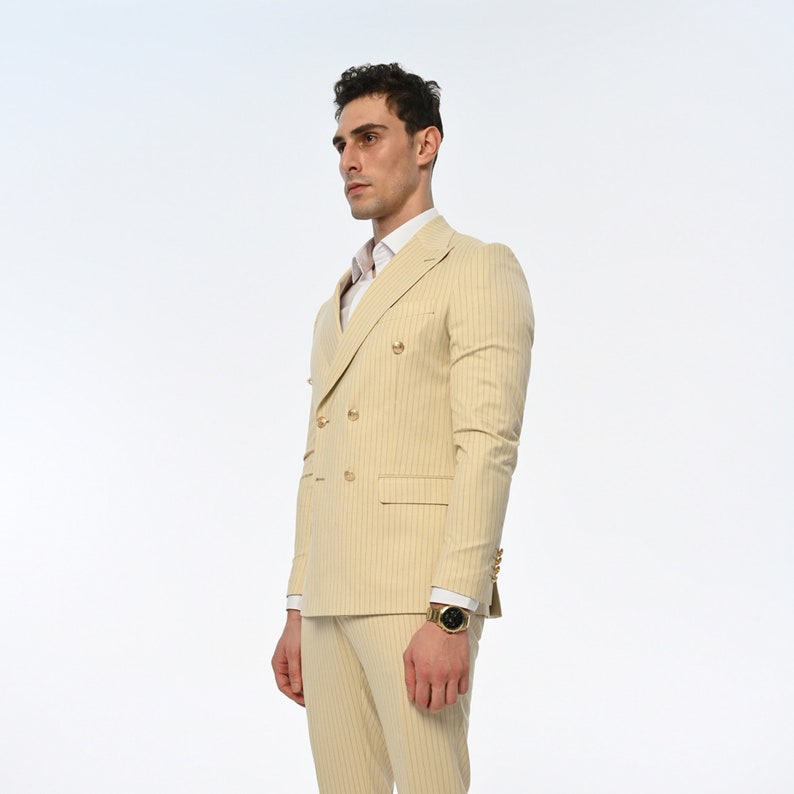 Men's suit consisting of cream, double-breasted, pointed wide collar, pin stripe, decorative gold buttons, slim fit trousers and jacket represents not only elegance but also sophisticated elegance with its cream colour and double-breasted design.