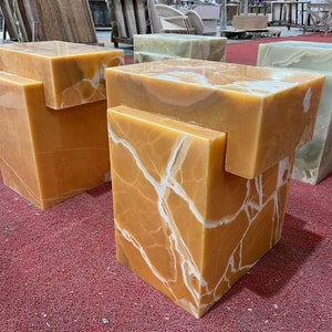 May include: Three decorative, square-shaped, orange onyx side tables with a unique, layered design. The tables have a natural, veined pattern and are made of polished stone.