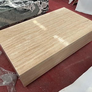 May include: A large, rectangular coffee table made of travertine stone. The table has a light beige color with a natural, striated pattern. The table is sitting on a red carpet.