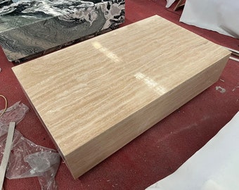 May include: A large, rectangular coffee table made of travertine stone. The table has a light beige color with a natural, striated pattern. The table is sitting on a red carpet.