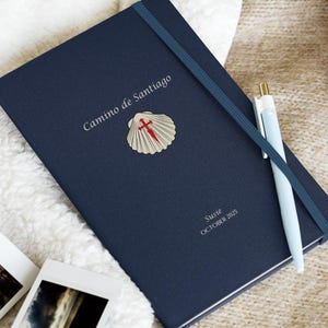 May include: A navy blue journal with the words "Camino de Santiago" and a shell emblem. A light blue pen rests on the journal, and a few small photos are nearby. The journal has a blue elastic band and a ribbon bookmark.