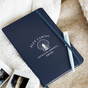 May include: A navy blue notebook with the text "Buen Camino" and a white scallop shell with a silhouette of two people walking. The text below the shell reads "Santiago de Compostela, Espana".