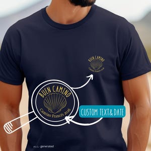 May include: A navy blue short-sleeve t-shirt featuring a small gold graphic on the chest. The graphic depicts a scallop shell with the text "BUEN CAMINO" and "Camino Francés 2026". A magnifying glass illustration highlights the design detail.