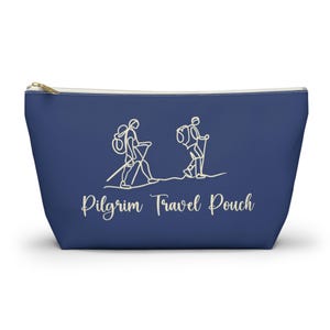 May include: A navy blue zippered pouch with a gold zipper and a white Pilgrim Travel Pouch design. The design features two line-drawn figures hiking with backpacks and walking sticks. The pouch is a travel accessory.