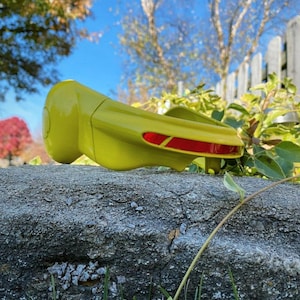 Peut inclure: Une visière en plastique jaune avec une bande rouge sur le dessus. La visière est posée sur un rocher avec de l'herbe verte en arrière-plan.