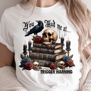 May include: White t-shirt with a black raven, skull, and books. The text "You had me at... Trigger Warning" is printed on the shirt.