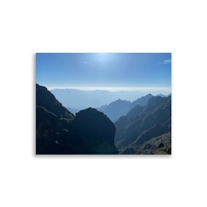 Könnte beinhalten: Ein Landschaftsdruck mit einer Bergkette unter einem strahlend blauen Himmel. Das Bild fängt den geschichteten Effekt der Berge ein, mit verschiedenen Blau- und Grüntönen, die ein Gefühl von Tiefe und Weite erzeugen.