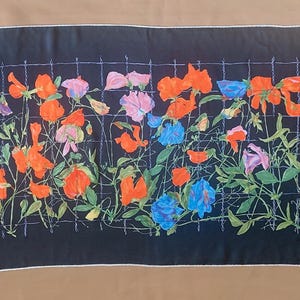 Puede incluir: Una bufanda larga y rectangular negra con un borde blanco. La bufanda presenta un diseño floral colorido con flores naranjas, rosas, moradas y azules, y hojas verdes. Las flores están dispuestas frente a una cuadrícula blanca.