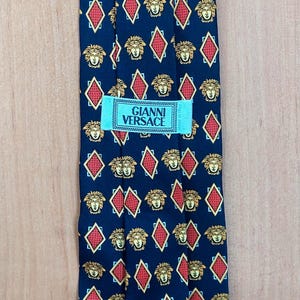 May include: Navy blue silk tie featuring a repeating pattern of gold Medusa heads and red diamond shapes. The tie has a label that reads "GIANNI VERSACE".