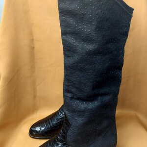 Puede incluir: Un par de botas negras hasta la rodilla con una caña texturizada de puntos y un pie de cuero brillante con patrón de reptil. Las botas tienen un tacón plano con un fino detalle metálico en tono dorado en la base del tacón y punta redondeada.