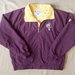 Può includere: Giacca Champion viola scuro con colletto giallo. La giacca ha una cerniera completa, polsini a coste e un piccolo logo ricamato sul petto. Un logo Champion è sulla manica.