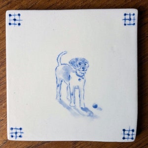 May include: A white ceramic tile featuring a blue and white illustration of a dog playing with a ball. The tile has a blue border with a grid pattern.