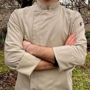 May include: A beige chef's jacket with long sleeves and a mandarin collar. The jacket is made of a smooth, wrinkle-resistant fabric.