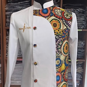 Könnte beinhalten: Weiße Kochjacke mit einem farbenfrohen Muster auf der rechten Seite. Das Muster zeigt verschiedene Kreise, Spiralen und Blumen in Rot-, Orange-, Gelb-, Blau- und Grüntönen. Die Jacke hat einen Stehkragen und vier schwarze Knöpfe auf der Vorderseite. Der Text "Muhammad INVAL" ist in Gold auf der linken Seite der Jacke gestickt.