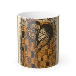 May include: White ceramic mug with a printed design of two figures in an embrace. The artwork uses brown, black, and gold tones, with a mosaic-style pattern. The mug has a standard handle and a white interior, suitable for hot drinks.