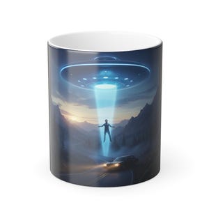 May include: A ceramic mug with a design depicting a UFO beaming up a person. The image shows a blue light beam, a flying saucer, a car on a road, and a mountain range in the background.