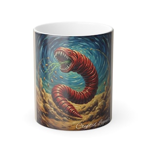 May include: A white ceramic mug featuring a detailed illustration of a red worm-like creature with a gaping mouth and sharp teeth. The creature is set against a blue and brown background. The text "Cryptid Legends" is printed on the mug.
