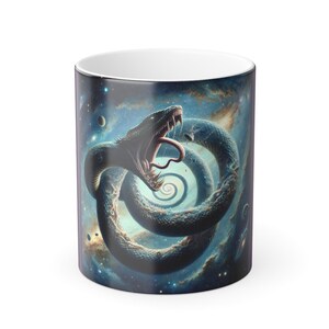 Könnte beinhalten: Weiße Keramik-Tasse mit einem Design einer aufgerollten Schlange mit offenem Maul, vor einem kosmischen Hintergrund aus Blau-, Grün- und Lilatönen. Die Zunge der Schlange ist sichtbar. Die Tasse hat einen weißen Innenraum.