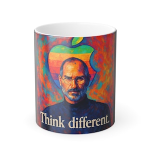 May include: A white ceramic mug featuring a colorful portrait of a man with glasses, set against a rainbow-colored apple logo. The words "Think different" are printed on the mug.