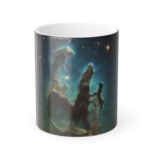 May include: White ceramic coffee mug featuring a vibrant image of the Pillars of Creation nebula. The design showcases a cosmic scene with blue, green, and brown hues, and a bright star. The mug has a glossy finish.