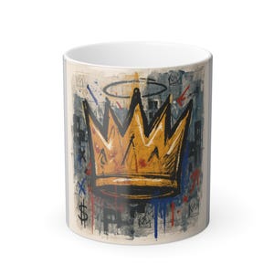 May include: White ceramic mug with a gold crown design and a halo, set against a backdrop of blue, red, and black graffiti-style art. The mug has a cylindrical shape and a white interior.