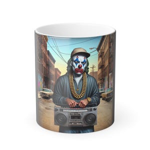 May include: White ceramic mug featuring a colorful illustration of a clown with a gold chain and a boombox. The clown is wearing a baseball cap and a gray jacket. The background depicts a city street scene.