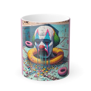 May include: White ceramic mug featuring a surreal illustration of a clown's head submerged in a pool. The clown has a painted face with red, blue, and white details, surrounded by a yellow inflatable ring and pink donuts. The background depicts a cracked pool.