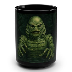 May include: Black ceramic mug featuring a green creature from the black lagoon. The creature has a textured, scaled appearance with sharp claws and a menacing expression. The background is a dark forest scene.