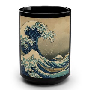 Op de afbeelding: Zwarte keramische mok met een print van Hokusai's "De Grote Golf van Kanagawa". Het kunstwerk toont een grote oceaangolf met boten en de berg Fuji op de achtergrond. De mok is cilindrisch en heeft een glanzende afwerking.
