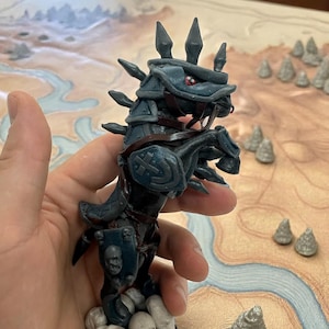 Peut inclure: Figurine détaillée d'une créature fantastique, peut-être un cheval ou un dragon, dans des tons bleu foncé et marron. La figurine a des pointes, des détails ressemblant à une armure et des yeux rouges. Elle est tenue devant une carte de jeu de table avec des éléments de terrain miniatures.
