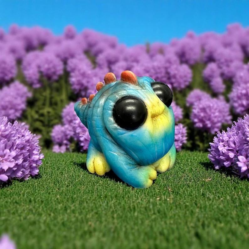 Op de afbeelding: Een klein, blauw en geel dinosaurusbeeldje met grote zwarte ogen. Het beeldje staat op een bed van groen gras met paarse bloemen op de achtergrond.