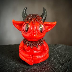 May include: A small, bright red creature figurine with large, glossy dark eyes, pointed ears, and two curved black horns. It features a textured brown collar of layered leaf-like shapes around its neck and a matching patch of brown hair between the horns.