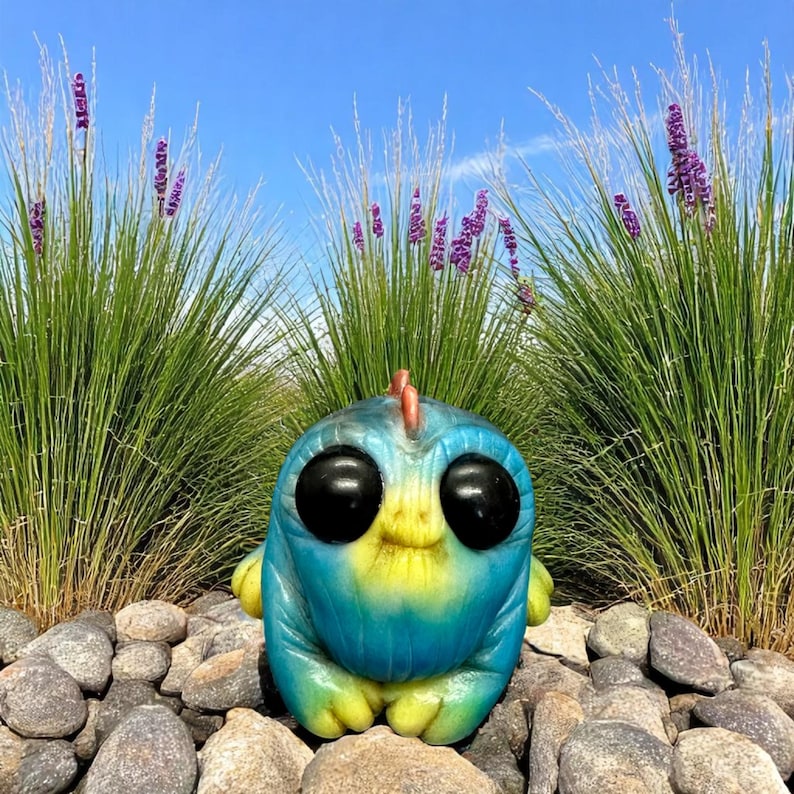 Op de afbeelding: Een handbeschilderd keramiek beeldje van een cartoonfiguur met grote ogen, in de kleuren blauw en geel. Het beeldje staat op een bed van grijze stenen.