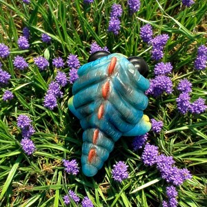 Op de afbeelding: Een blauw en rood dinosaurus speelgoed met een stekelige rug en zwarte ogen. Het speelgoed zit in een bed van paarse bloemen.