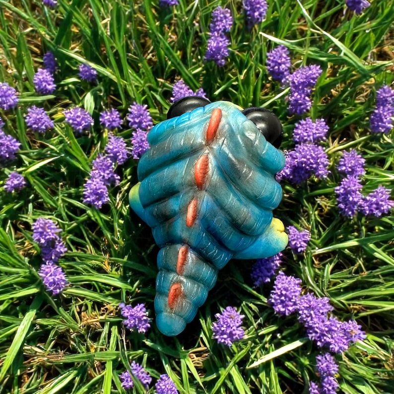 Op de afbeelding: Een blauw en rood dinosaurus speelgoed met een stekelige rug en zwarte ogen. Het speelgoed zit in een bed van paarse bloemen.
