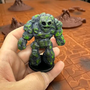 Puede incluir: Figura en miniatura de fantasía de un golem de piedra con detalles verdes tipo musgo. La figura tiene una cara con forma de calavera y está montada sobre una base negra. El golem se sostiene en una mano, con un fondo borroso de terreno.