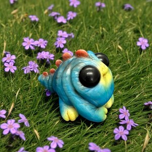 Op de afbeelding: Een klein, handgemaakt schepsel in blauw en geel met grote zwarte ogen en een stekelige staart. Het schepsel zit op een bed van groen gras en paarse bloemen.