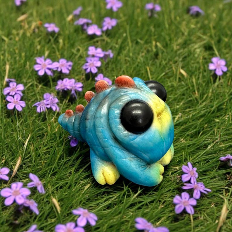 Op de afbeelding: Een klein, handgemaakt schepsel in blauw en geel met grote zwarte ogen en een stekelige staart. Het schepsel zit op een bed van groen gras en paarse bloemen.