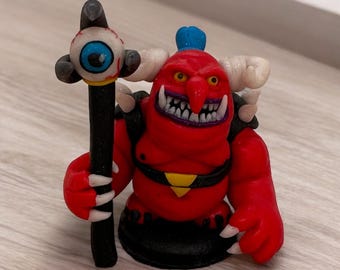 Könnte beinhalten: Eine rote und schwarze Dämonenfigur, die einen Stab mit einem großen Auge oben hält. Der Dämon hat eine große Nase, Hörner und ein zahniges Grinsen.
