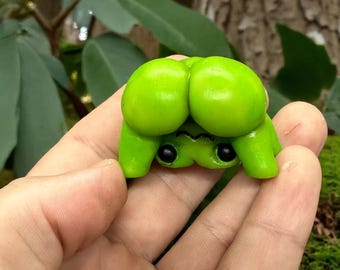 May include: A handmade, lime green frog figurine with large, round eyes and a whimsical design. The frog is held in a hand, showcasing its bright color and unique shape. The figurine is a playful, artistic creation.