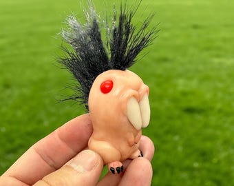 Könnte beinhalten: Eine skurrile Figur mit einem hellpfirsichfarbenen Körper, großen weißen Zähnen und einem roten Auge. Sie hat ein schwarzes und silbernes, stacheliges, haarähnliches Merkmal oben. Die Figur hat kleine schwarze Füße und wird in einer Hand vor einem grünen Hintergrund gehalten.