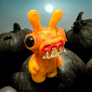 May include: A whimsical orange creature figurine with large black eyes, a wide mouth revealing sharp teeth, and long ears. The figurine is set against a backdrop of dark pumpkins and a moon, creating a Halloween theme.