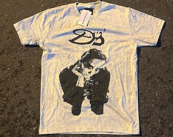 May include: A light gray t-shirt with a black and white graphic print of a person sitting down. The graphic includes the text "25".