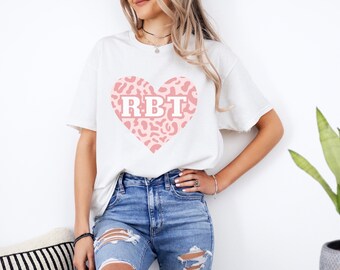 May include: White t-shirt featuring a pink leopard print heart with the letters "RBT" in white. The shirt is paired with blue jeans. The overall style is casual and relaxed.