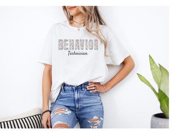 May include: White t-shirt with the words "BEHAVIOR Technician" in a floral design. The model is wearing blue jeans. The shirt is a casual, everyday wear item.