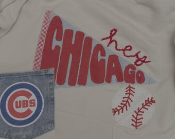 May include: Cream hoodie with a Chicago theme. It has a red and blue pennant with "CHICAGO" and "hey GO" embroidered, a baseball patch, and a denim pocket with a Cubs logo.