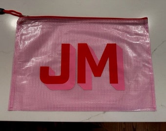 May include: A pink, zippered pouch with the letters "JM" in red and pink. The pouch has a red zipper and a black pull tab. The material appears to be a transparent, grid-like plastic.