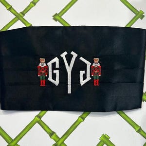 May include: Black decorative sash with white embroidered initials and two embroidered nutcracker figures in red, green, and gold. The sash is rectangular and has a pleated design.