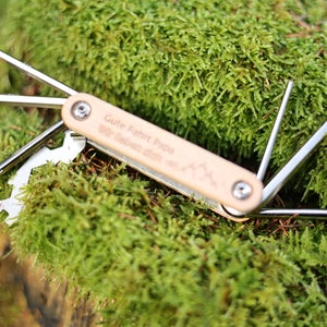 Op de afbeelding: Een multitool met een houten handvat gegraveerd met de tekst "Gute Fahrt Papa" en een bergontwerp. De tool heeft meerdere hulpstukken, waaronder een schroevendraaier, een flesopener en een sleutel.