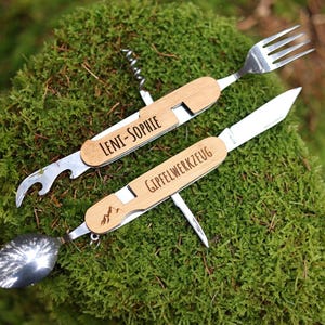 Könnte beinhalten: Zwei Multitools mit Holzgriffen und Klingen aus Edelstahl. Das obere Werkzeug hat einen Flaschenöffner, einen Korkenzieher, eine Gabel und den Text "LENI-SOPHIE". Das untere Werkzeug hat einen Löffel, ein Messer und den Text "GIPFELWERKZEUG".